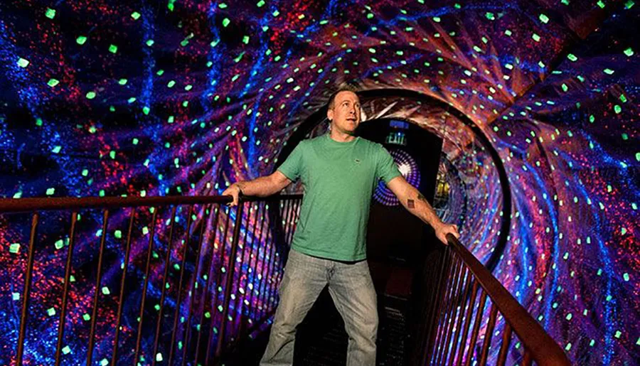 A person stands on a bridge inside a dazzling, tunnel-like space with walls covered in colorful, luminous patterns.