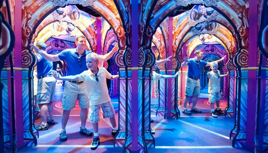A smiling adult and child are posing with outstretched arms in a vibrant and colorful mirror maze.