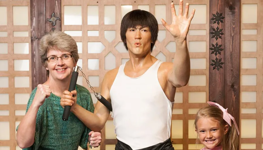 Two people and a life-like figure that resembles a martial arts performer are posing for a picture with smiles and a nunchaku prop.