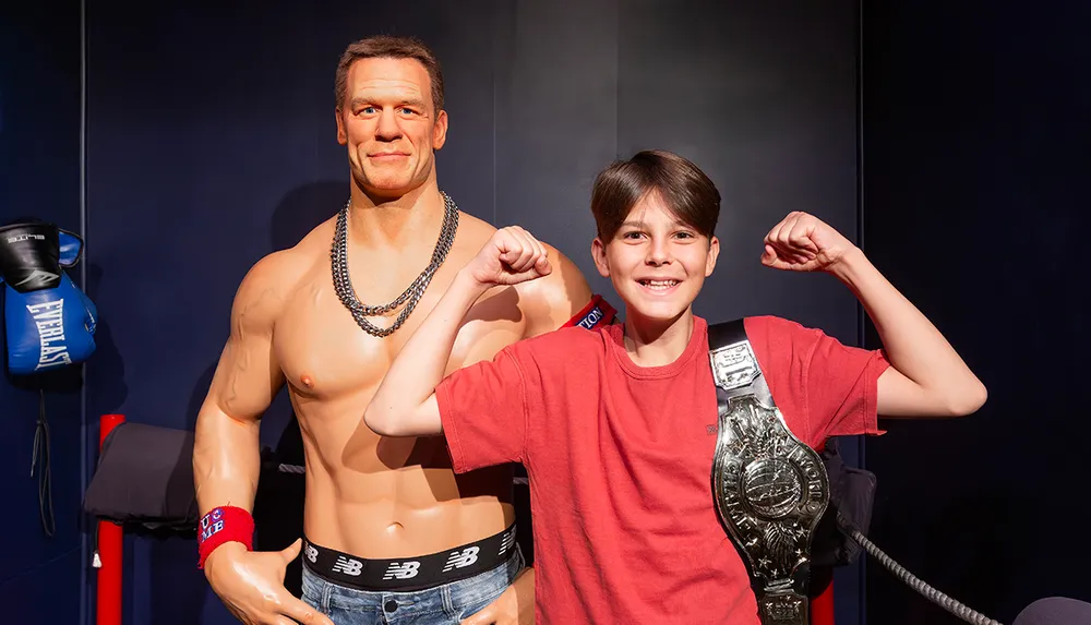 A child is posing with a muscular wax figure while flexing their arms and wearing a wrestling championship belt