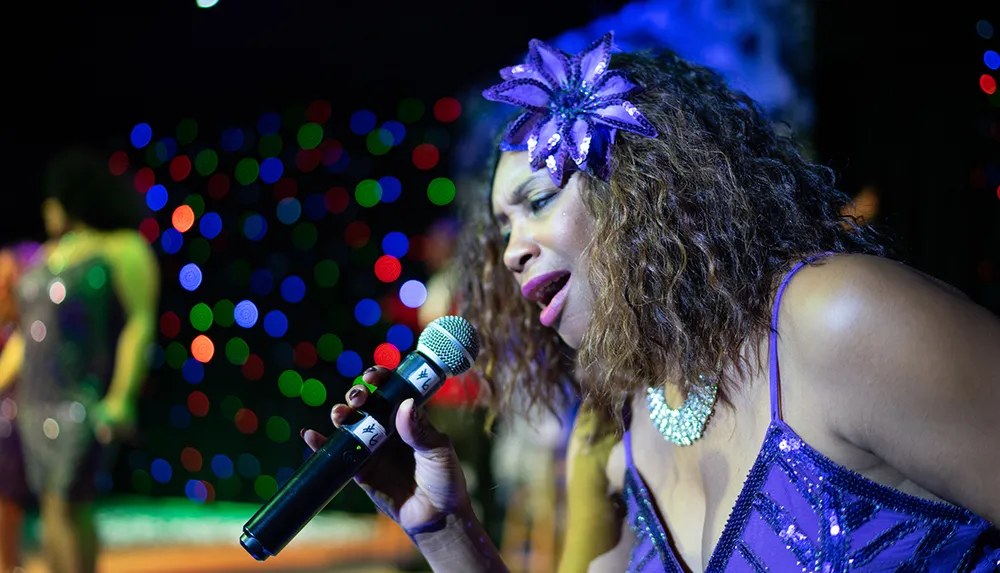 A woman adorned in a sparkly purple outfit passionately sings into a microphone against a backdrop of colorful bokeh lights