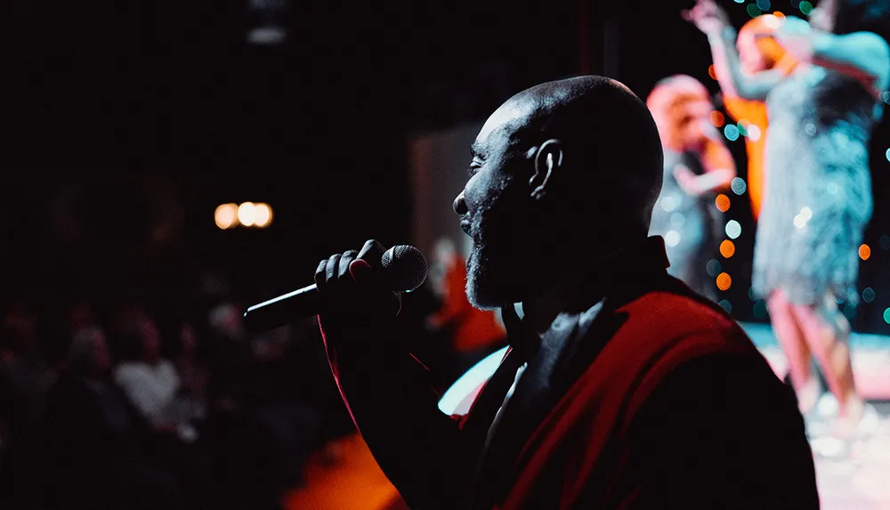 A performer is singing into a microphone on stage with an audience in the background and another performer visible behind him