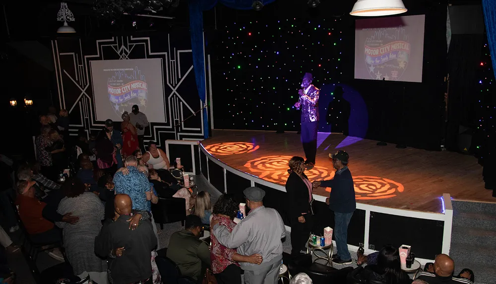A performer is on stage at a Motor City Musical event while an engaged audience watches from their seats around the venue
