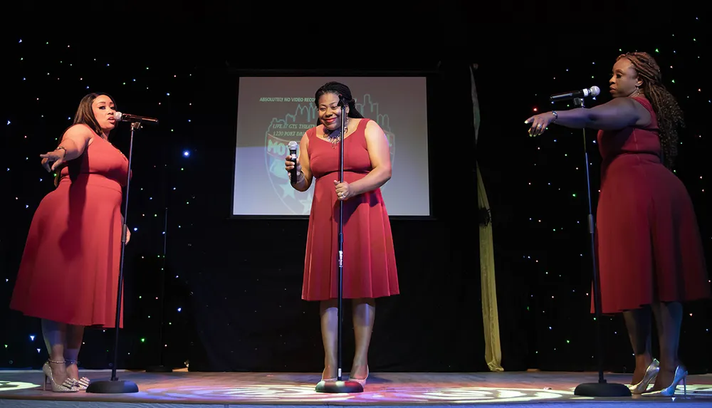 Three women in matching red dresses are performing on stage each singing into a microphone with a backdrop of a lit screen and twinkling lights