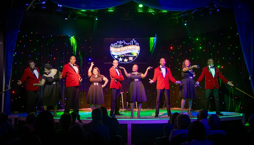 A group of performers in formal attire is on stage singing and entertaining an audience under colorful stage lighting and a backdrop that reads Motor City Musical