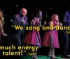 A group of five singers is performing on stage with colorful stage lights in the background