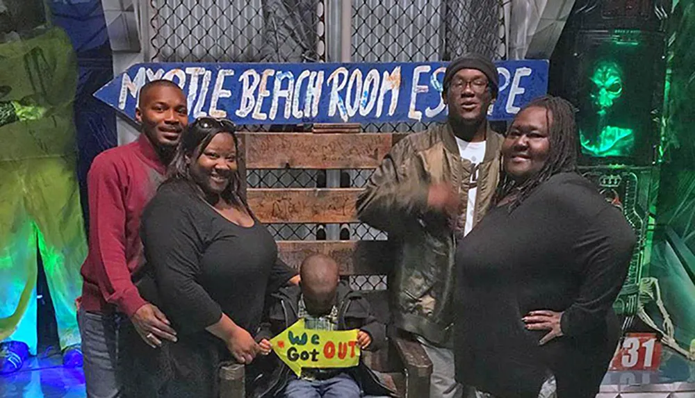 A group of people is celebrating their successful escape from a room escape challenge at Myrtle Beach Room Escape with a child holding a We Got Out sign