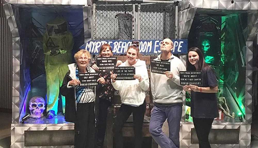 A group of people is posing with playful mugshot placards in front of a horror-themed backdrop likely commemorating their escape room experience