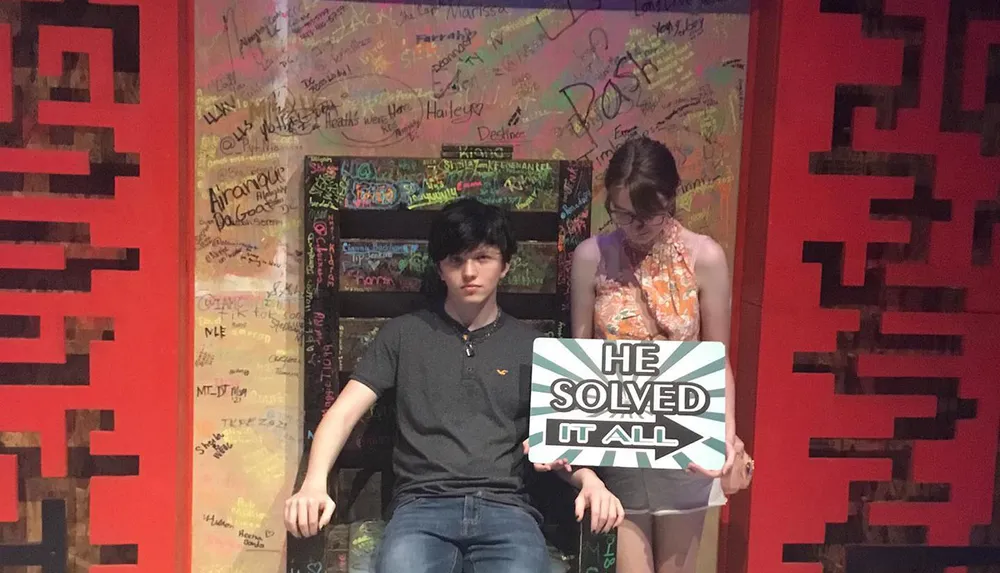 A young man and woman are seated on a throne-like chair with walls covered in graffiti and the woman is holding a sign that says HE SOLVED IT ALL