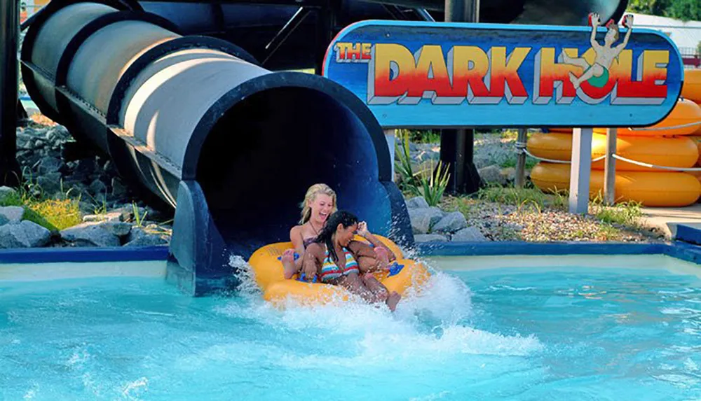 Two people are gleefully exiting a water slide called The Dark Hole on a yellow double inner tube at a water park
