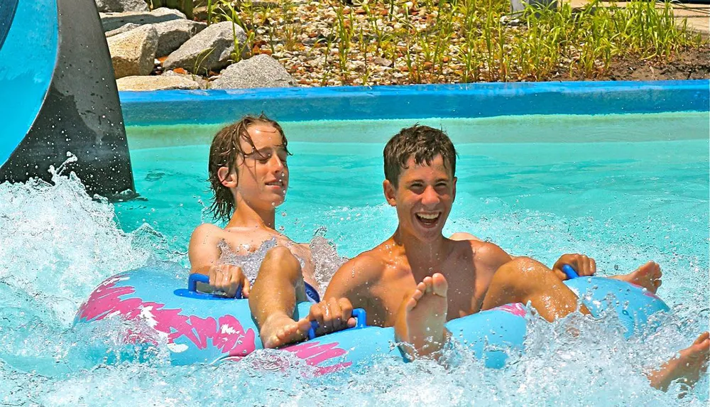 Two individuals are joyfully riding on an inflatable tube in a water slide splashing into the water with big smiles on their faces