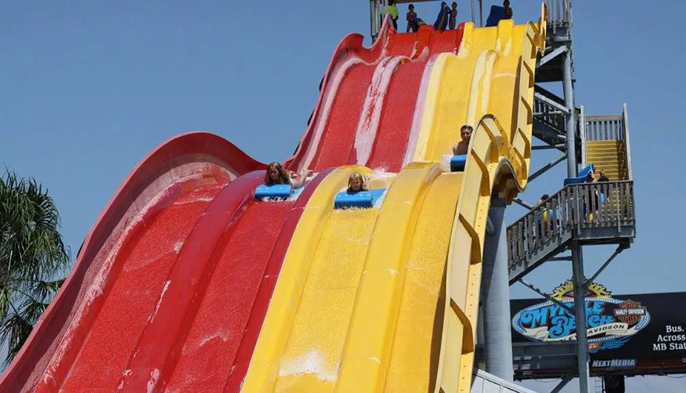 This image shows people sliding down a colorful water slide at an amusement park with some waiting at the top for their turn