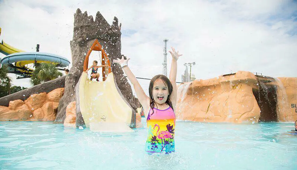 A joyful child is raising her arms in excitement in a water park with a small water slide and splashing water in the background