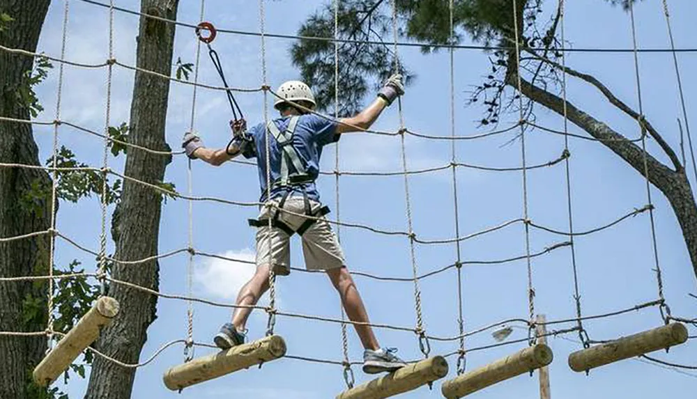 A person wearing a helmet and harness is balancing on logs suspended by ropes as part of an outdoor high ropes adventure course