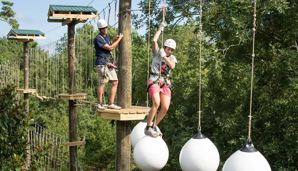 Two individuals wearing helmets are engaging in a rope course adventure among trees with one person traversing white spherical obstacles