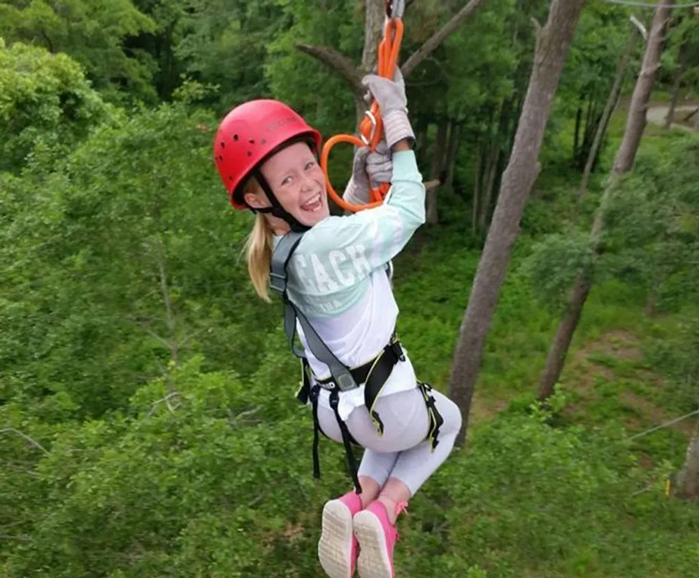 A young girl is smiling joyfully while zip-lining amongst trees wearing a helmet and harness for safety
