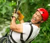 A person wearing a red helmet and a harness is enthusiastically zip-lining amidst green foliage