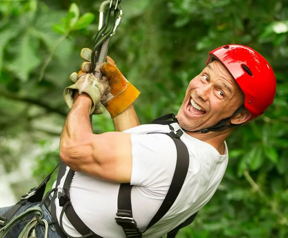 A person wearing a red helmet and a harness is enthusiastically zip-lining amidst green foliage