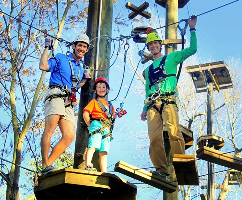 Three people are smiling and posing on a high ropes adventure course among trees equipped with helmets and safety harnesses