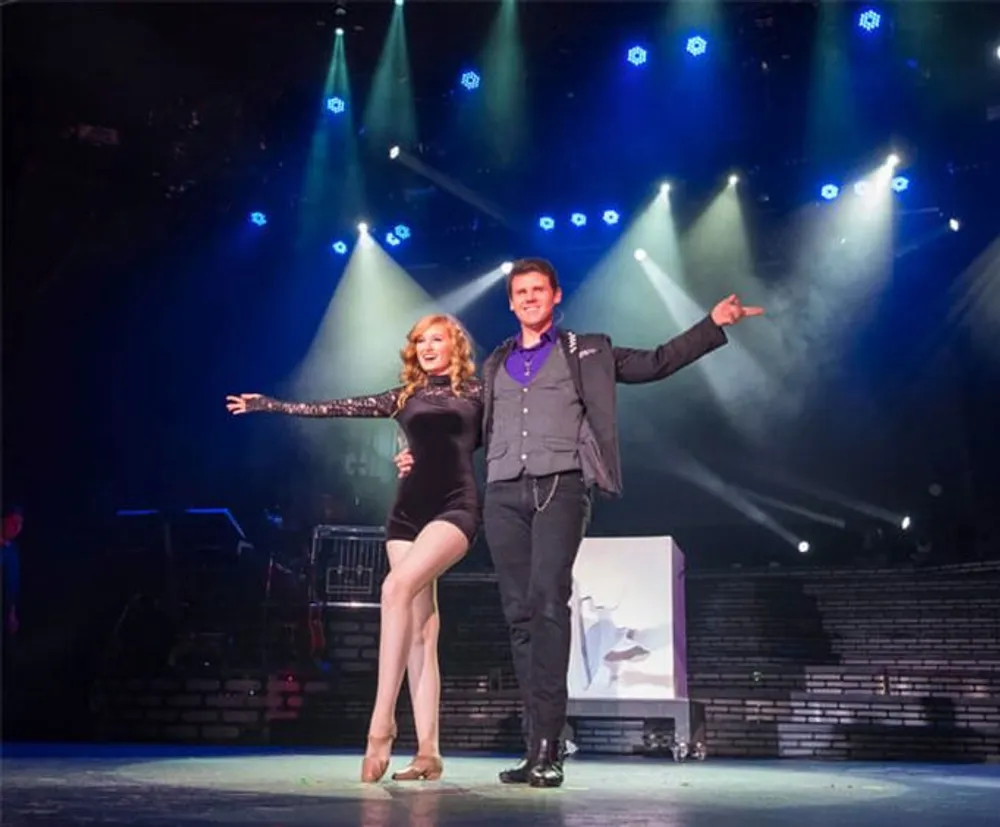 A man and a woman are posing confidently on a stage with bright stage lights shining from above