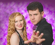 A smiling woman with red hair and a man in a dark suit striking a dramatic pose with an outstretched hand in front of a purple bokeh background.