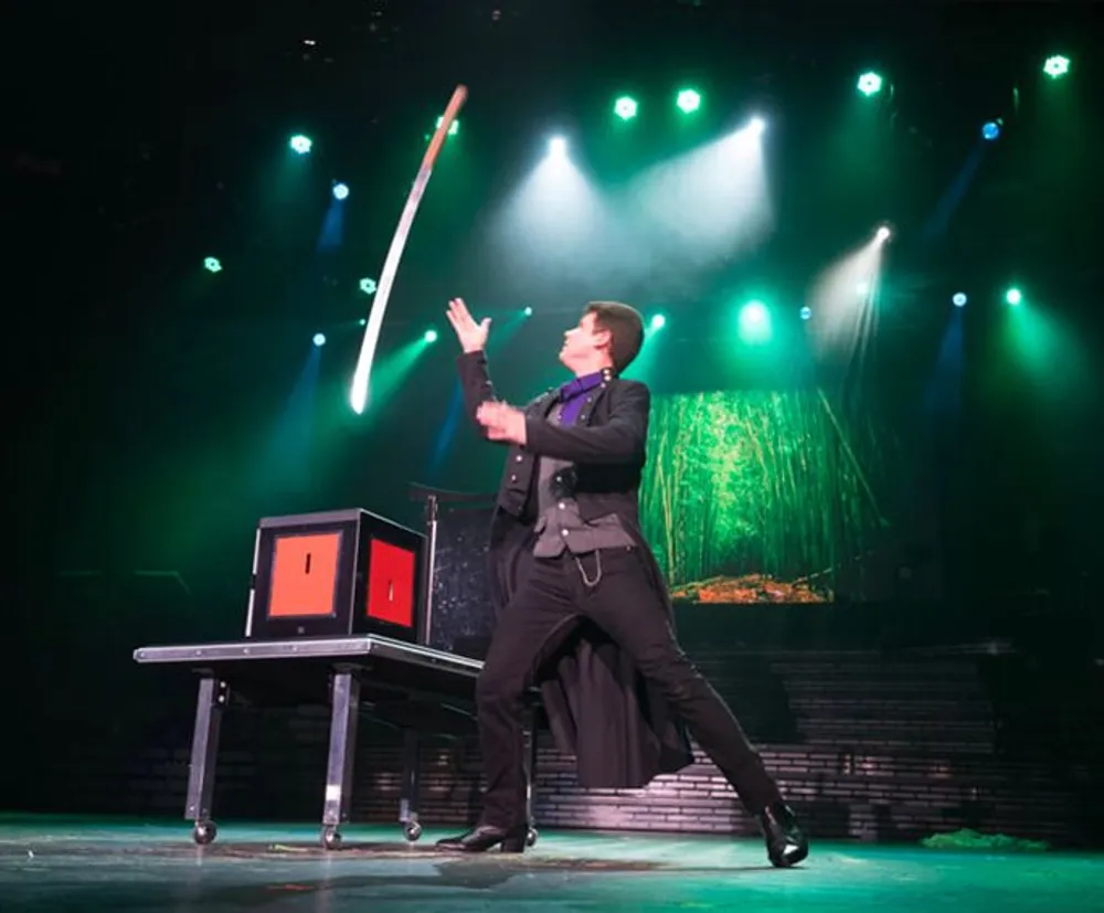 A magician is on stage performing a trick throwing a baton in the air with a spotlight and stage lights illuminating the scene