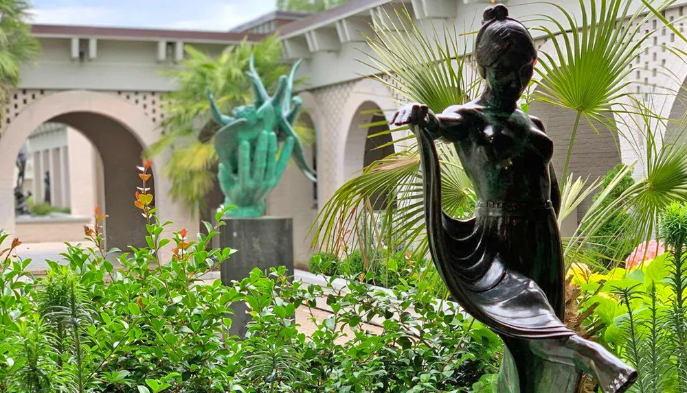A bronze statue of a dancing figure is foregrounded against a lush garden with another statue of a hand in the background