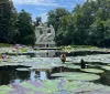 The image shows a tranquil pond with vibrant water lilies in the foreground and an imposing white sculpture of figures in the background all set amidst lush greenery