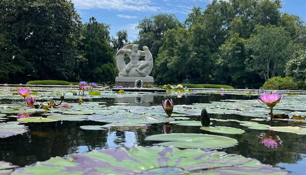 The image shows a tranquil pond with vibrant water lilies in the foreground and an imposing white sculpture of figures in the background all set amidst lush greenery