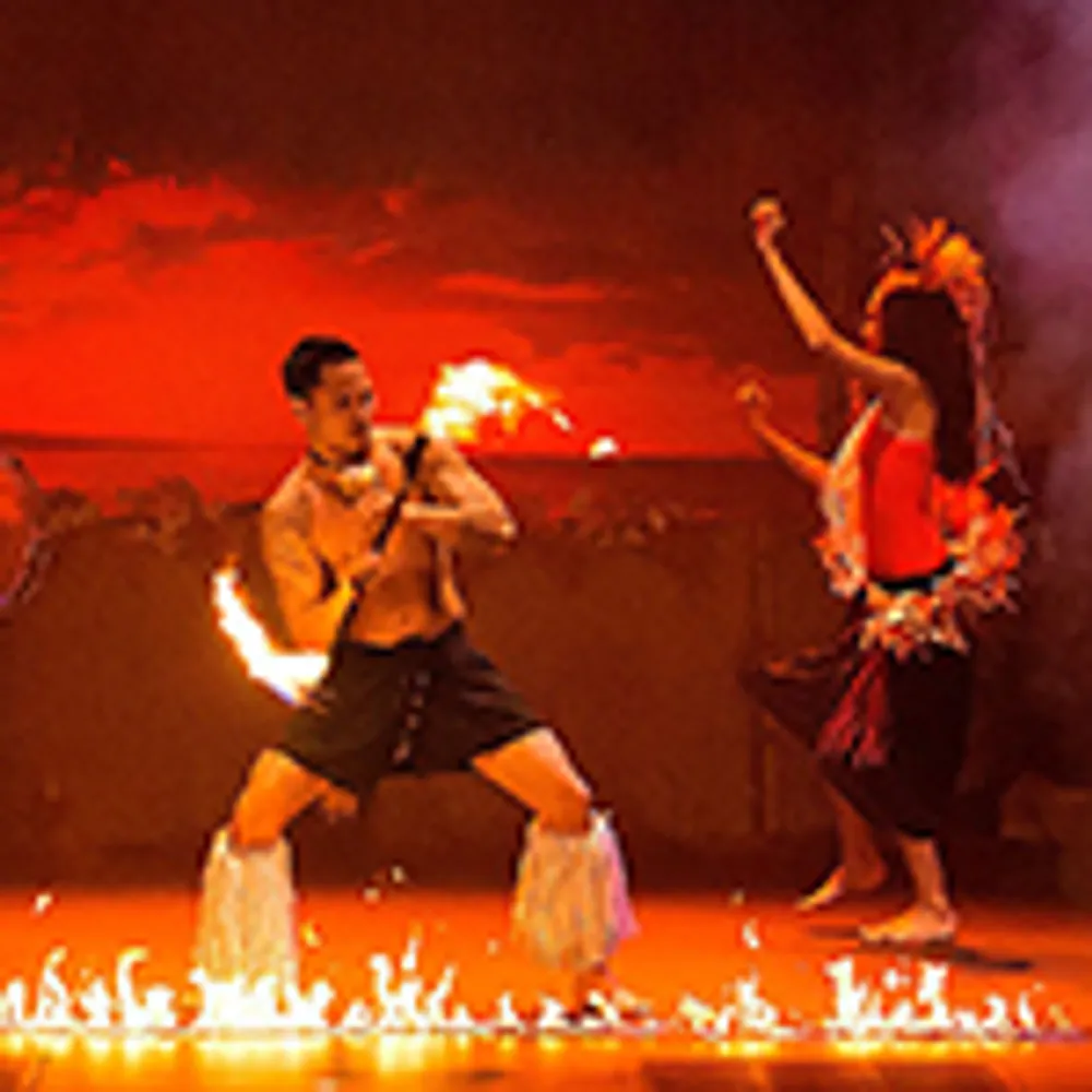 The image shows performers engaged in a vibrant fire dance against a dramatic red-lit background