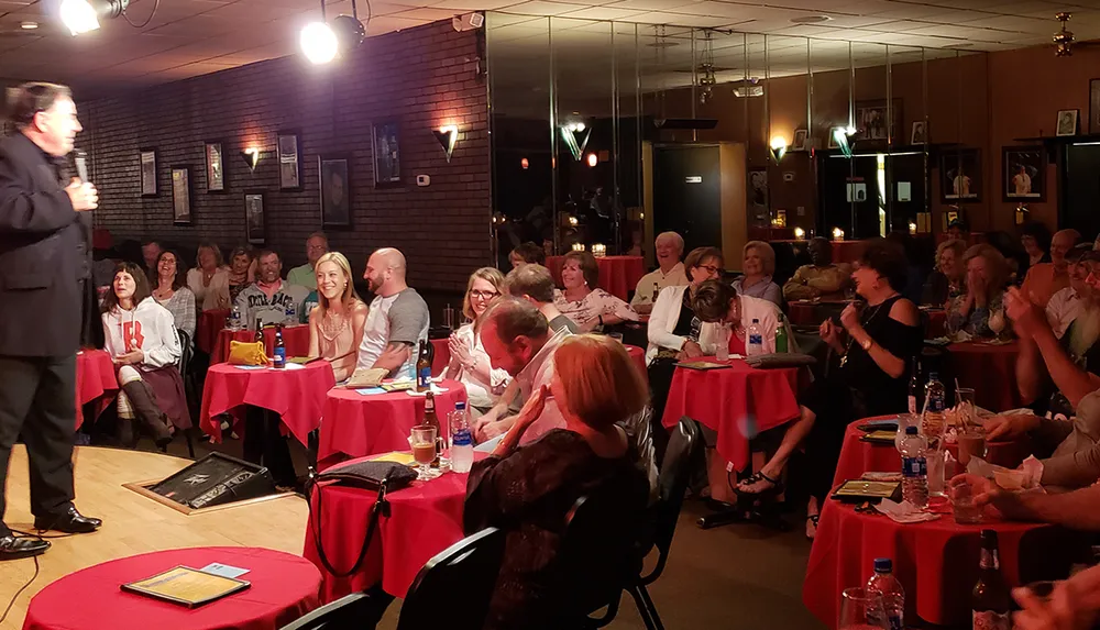 A comedian is performing on stage in front of an audience seated at tables with red cloths eliciting smiles and laughter in a cozy dimly-lit comedy club