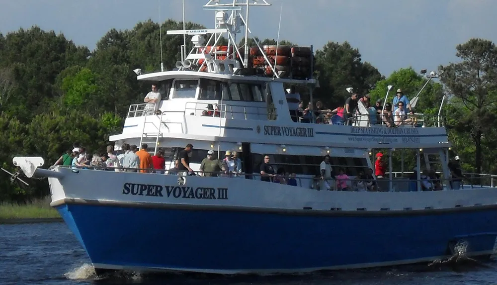 A large group of passengers is aboard the SUPER VOYAGER III a blue and white boat designated for deep sea fishing and dolphin watching cruising near a shoreline