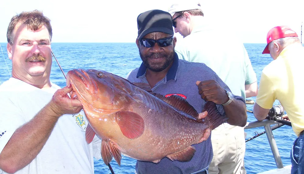 Two men are holding a large fish with a smile on a boat surrounded by other people who are also fishing at sea