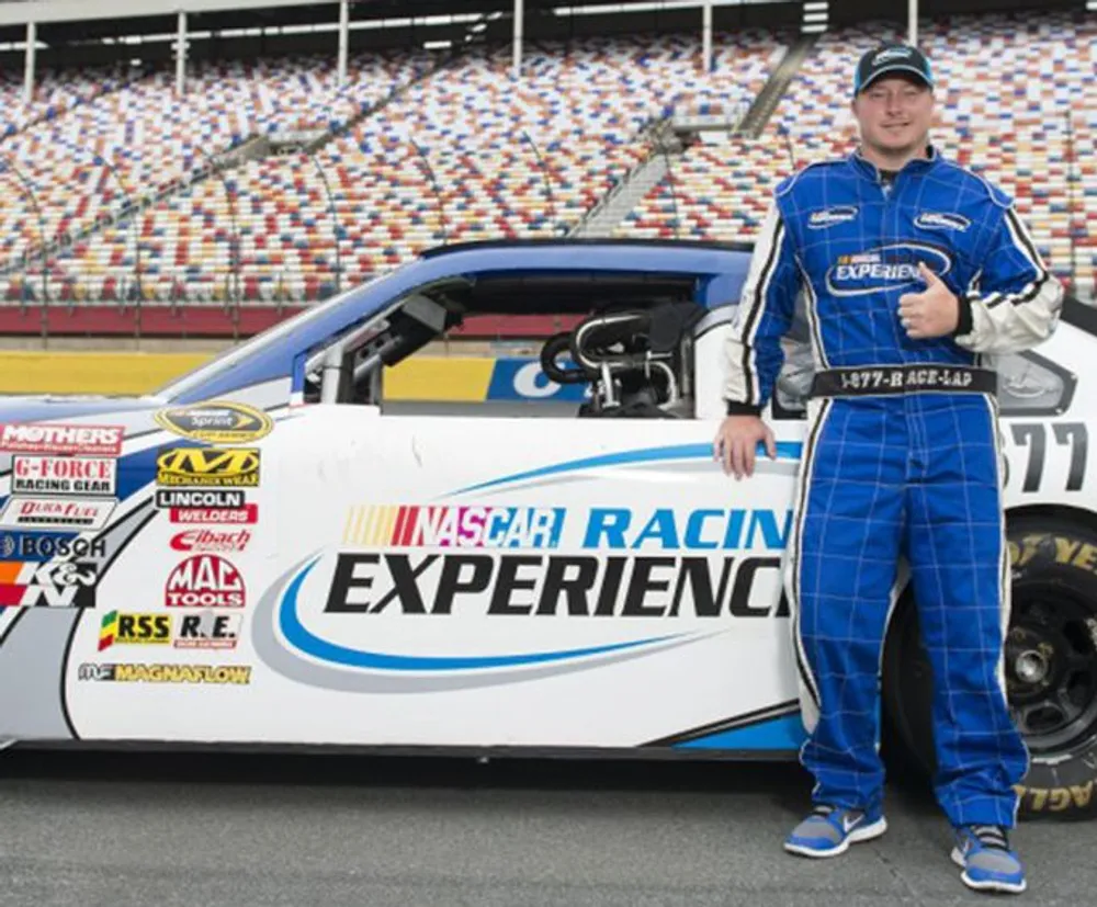 A person in a blue racing suit is giving a thumbs-up next to a white NASCAR race car with the text NASCAR Racing Experience on its side at a speedway with empty stands in the background
