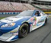 A person wearing a racing helmet is sitting inside a NASCAR race car adorned with sponsor logos parked on a track with grandstands in the background