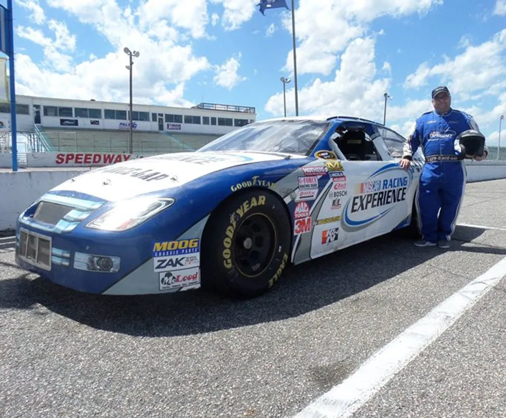 A person in a racing suit stands beside a blue and white NASCAR race car at a speedway