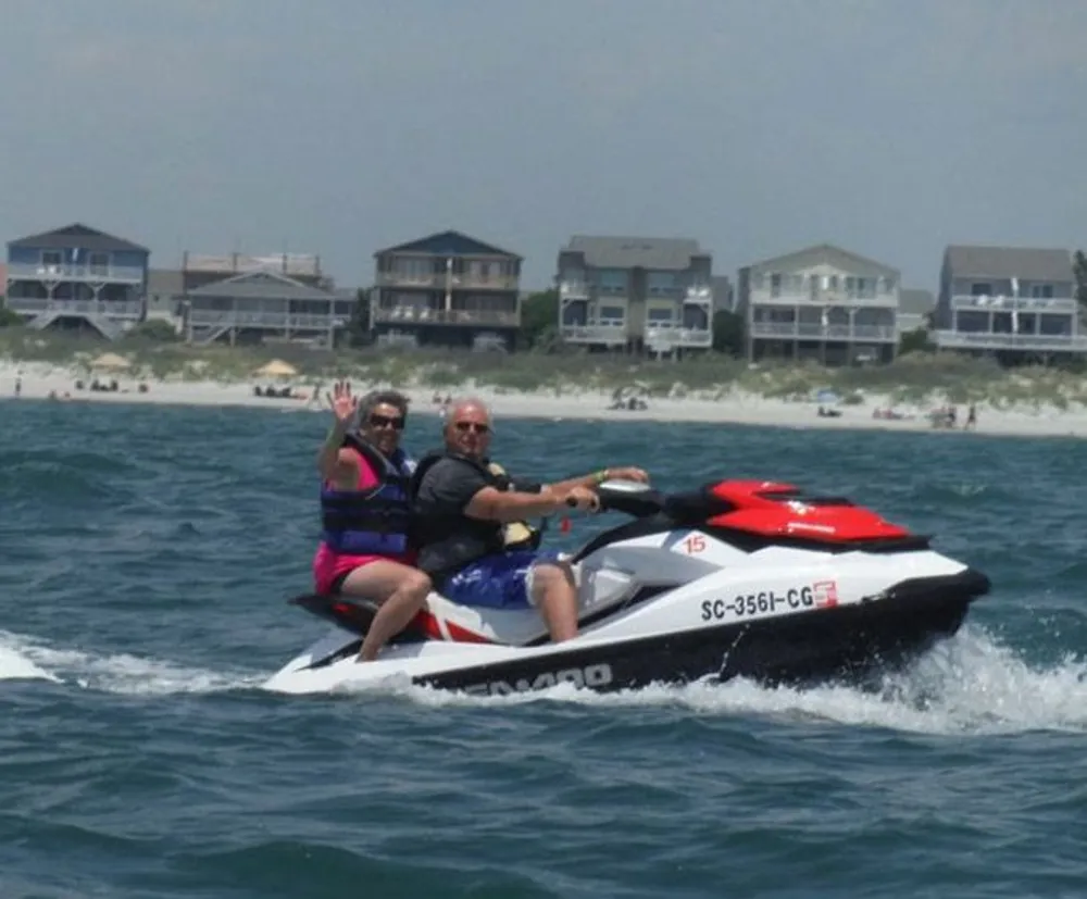 Two people a woman and a man are riding a jet ski near the coastline with beach houses in the background and the woman is waving at the camera