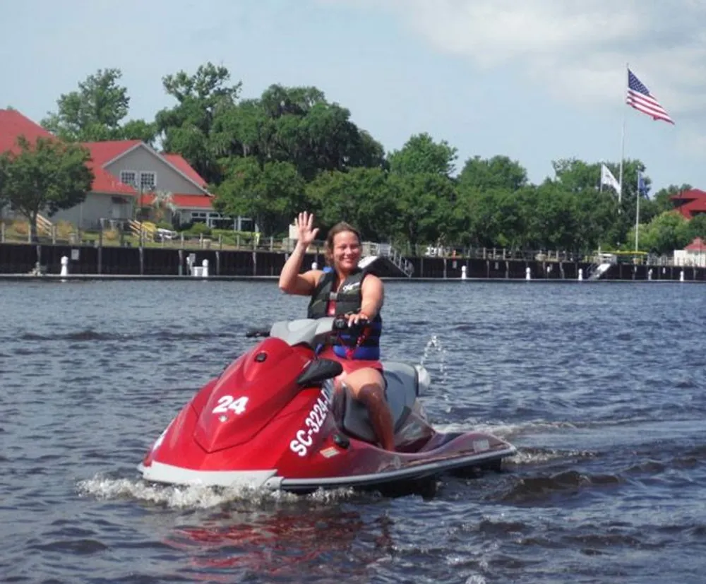 A person is waving while riding a red jet ski on a calm waterway with buildings and an American flag in the background