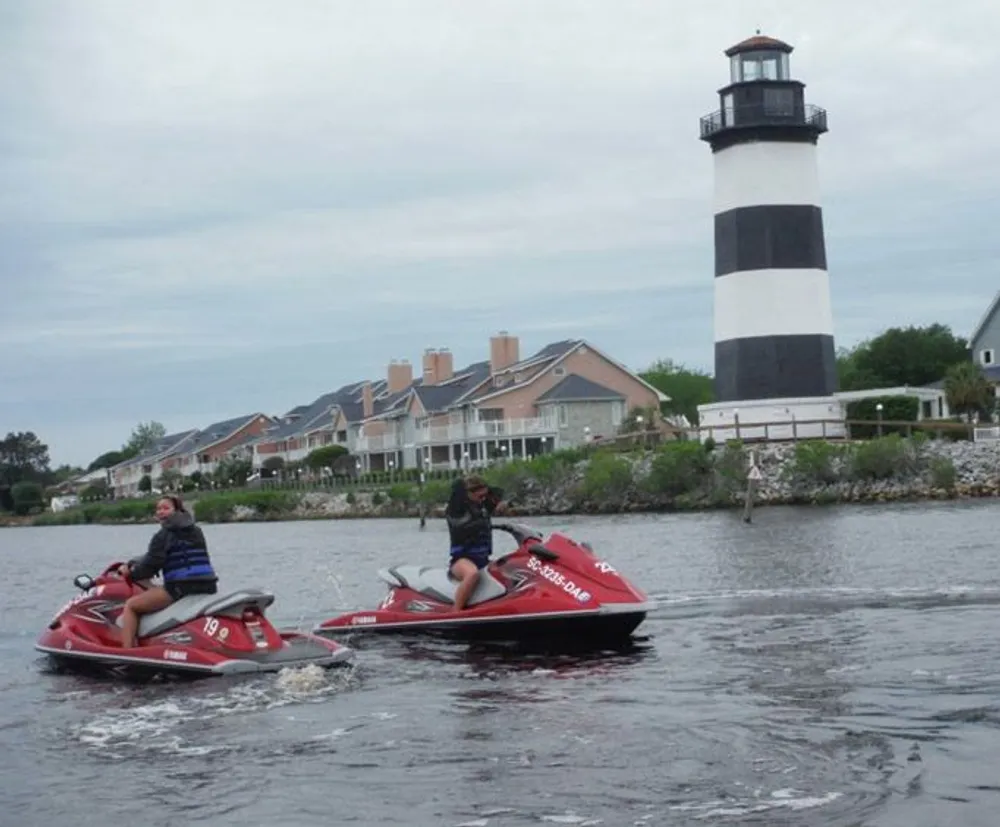 Two people are riding jet skis near a lighthouse with houses in the background