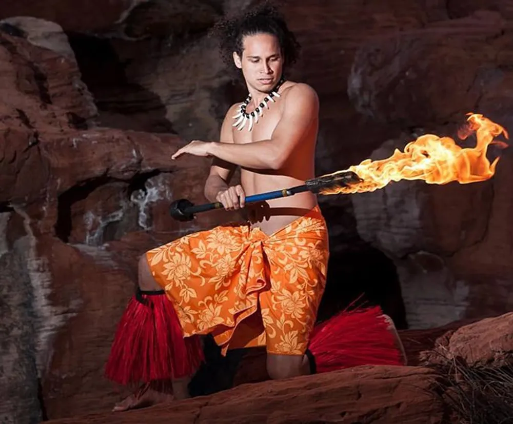 A person is performing a traditional fire dance in front of a rocky backdrop wearing cultural attire consisting of an orange-patterned sarong and a necklace and wielding a flaming staff