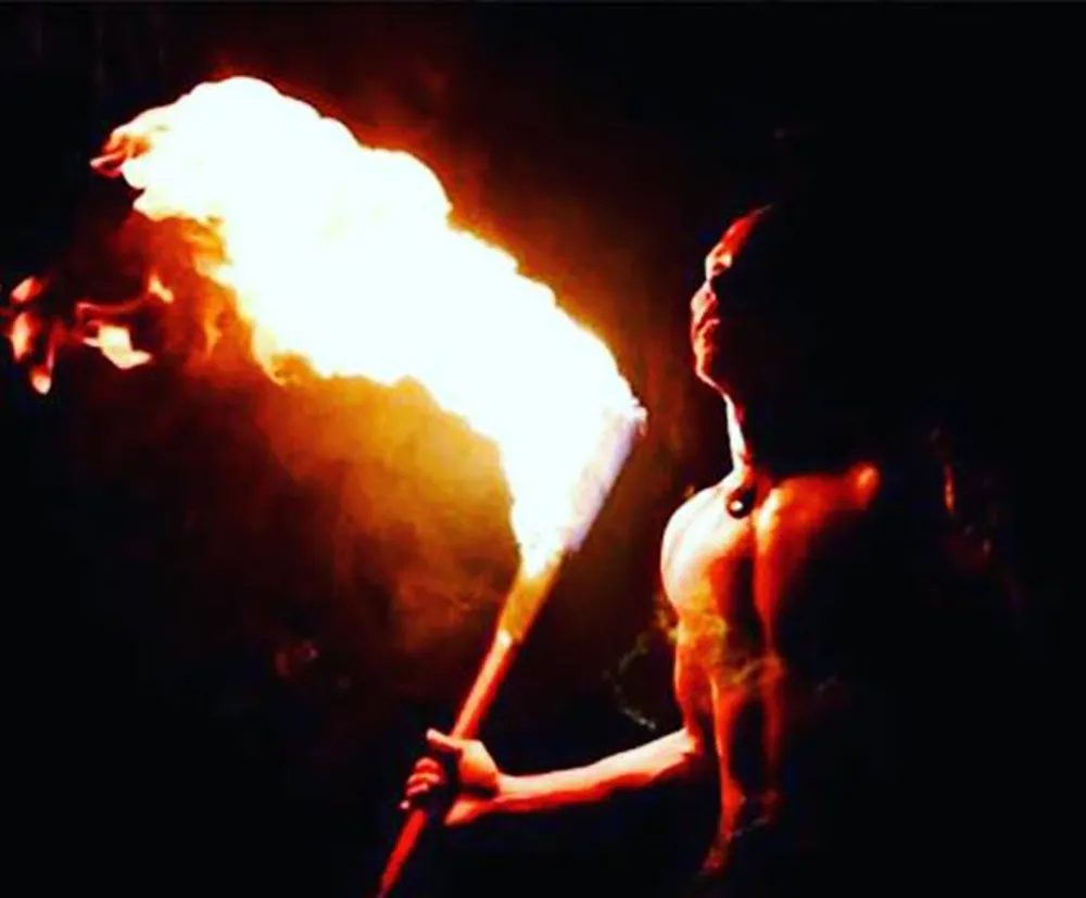 This image depicts a person performing fire breathing exhaling a large flame from their mouth while holding a torch