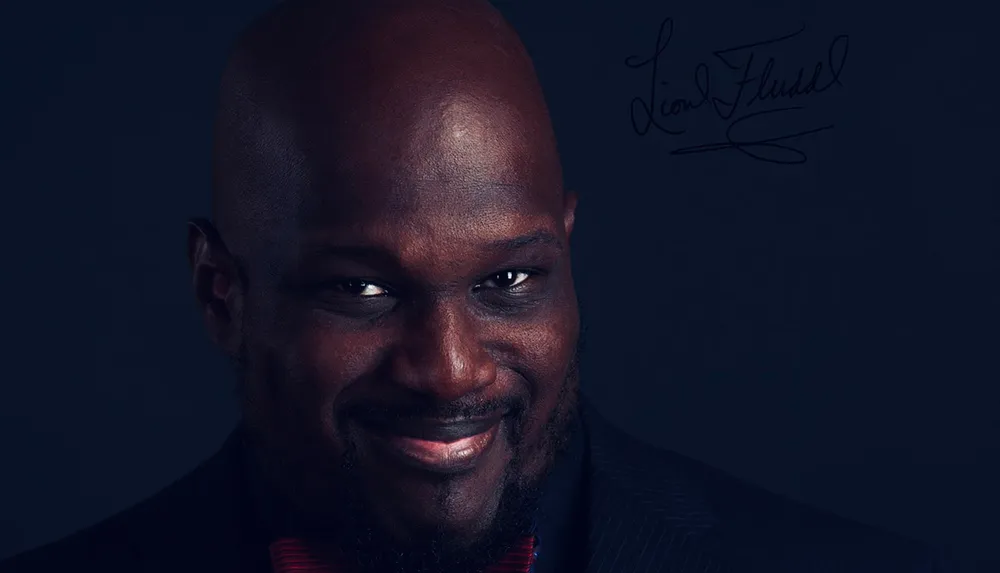 The image features a smiling bald man with a beard wearing a dark jacket with a red striped tie set against a dark background with a signature overlay on the right side