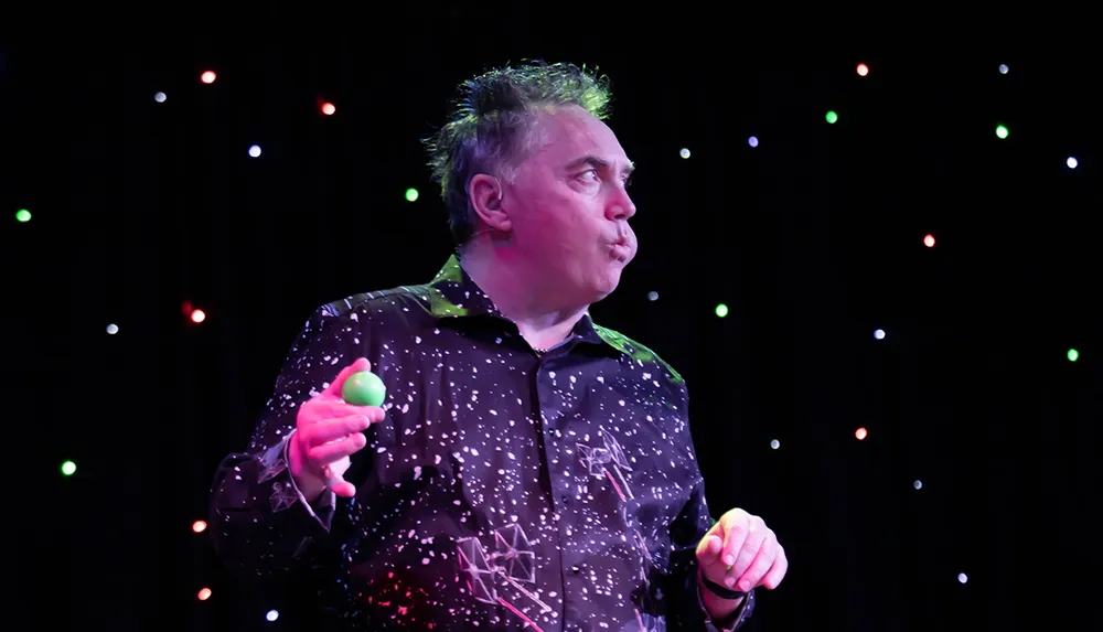 A performer stands on stage seemingly mid-act with colorful lights in the background wearing a patterned shirt splattered with white and holding a green shaker in one hand