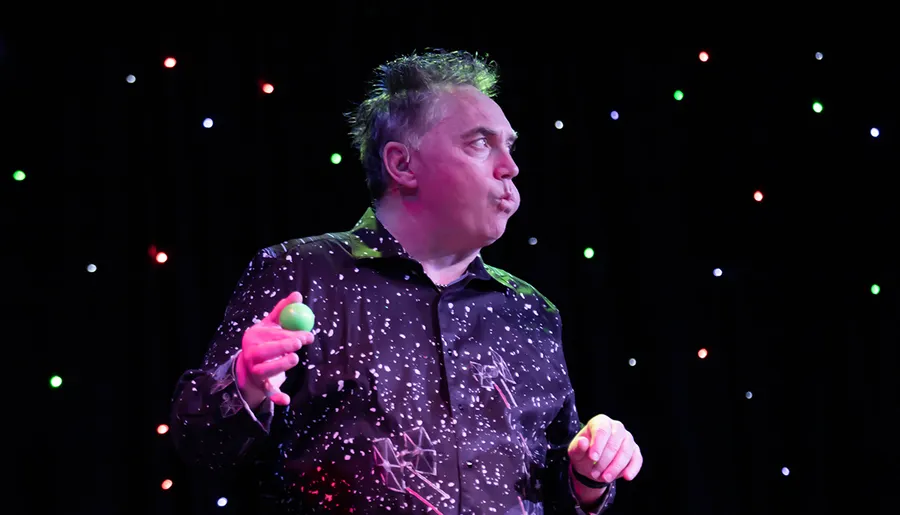 A performer stands on stage, seemingly mid-act, with colorful lights in the background, wearing a patterned shirt splattered with white, and holding a green shaker in one hand.