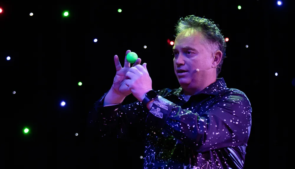 A person is performing on stage with a green ball in hand wearing a headset microphone against a backdrop of colorful lights