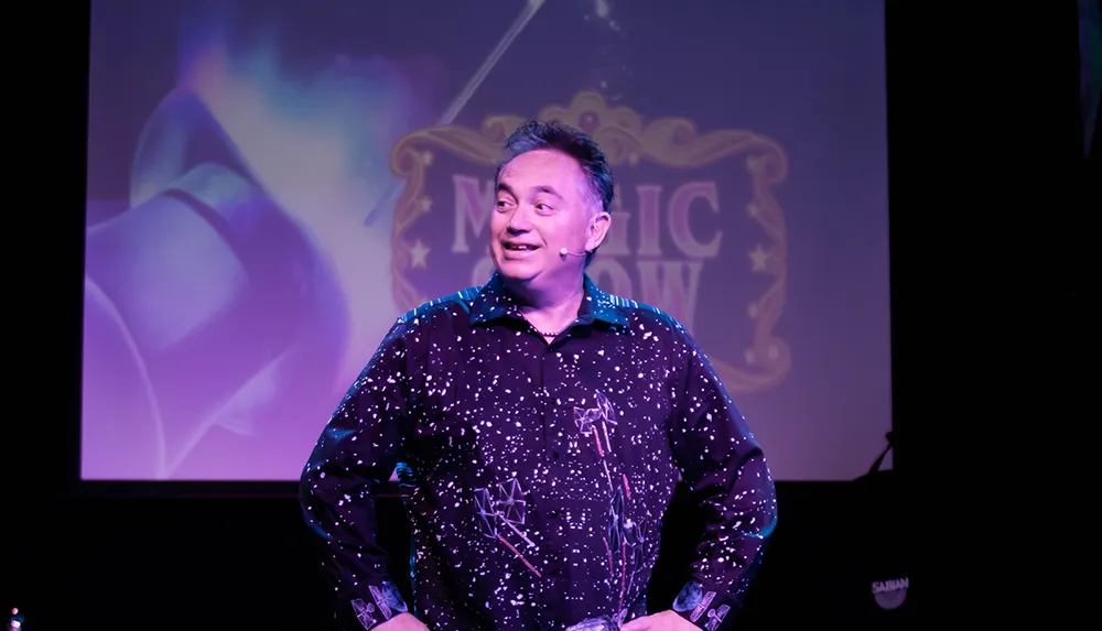 A person in a star-patterned shirt with a headset microphone is standing on stage with a backdrop that reads MAGIC SHOW