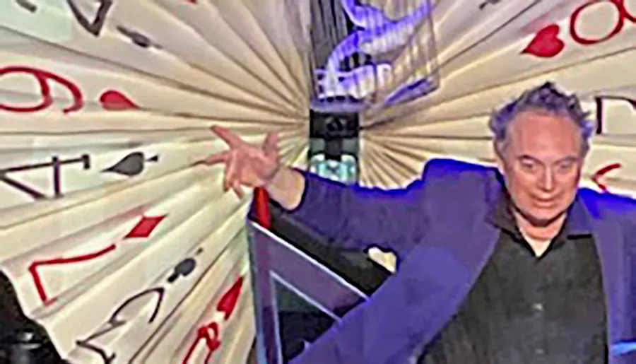 A person stands in front of a large, fanned-out display of oversized playing cards, striking an expressive pose.