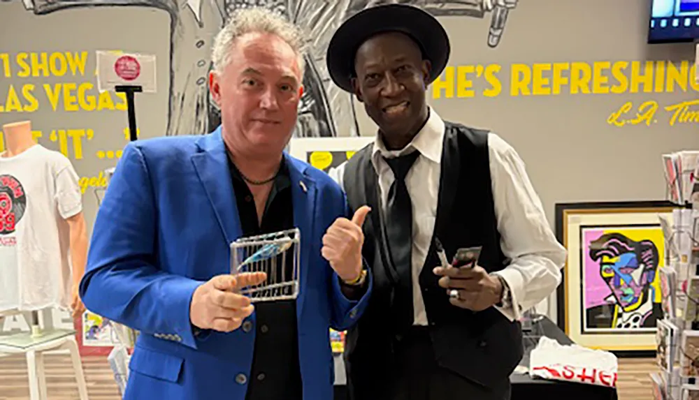Two men dressed in suits pose together indoors with one holding an award and the other giving a thumbs-up gesture