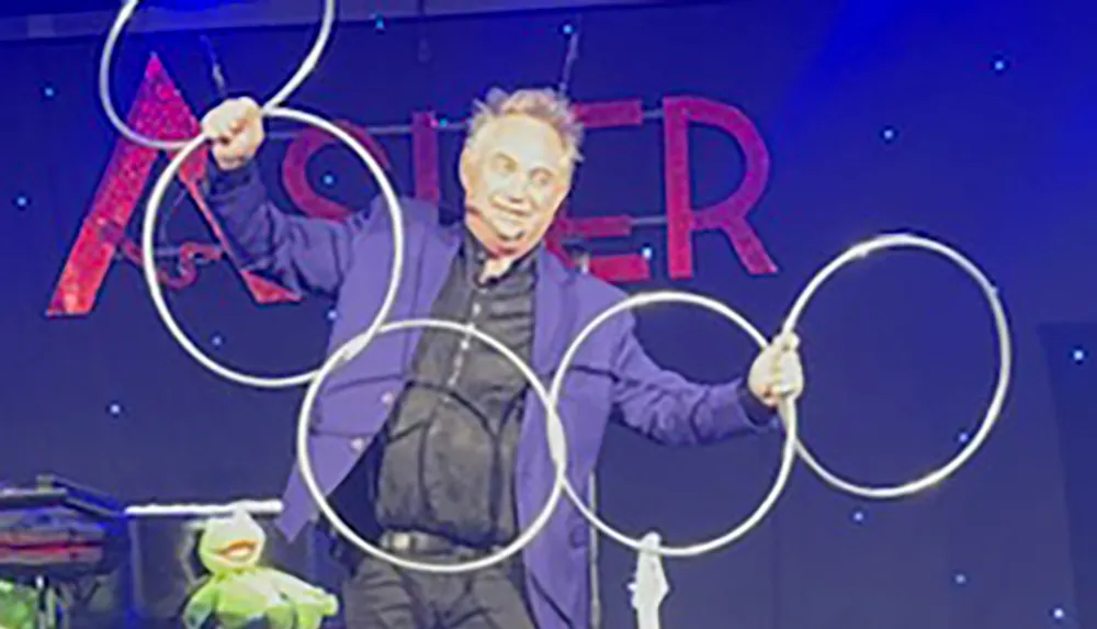 A performer is on stage holding multiple interlocking rings with a puppet resembling Kermit the Frog on the table