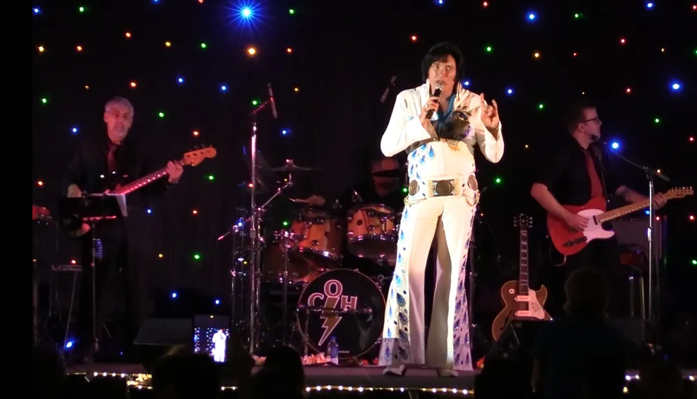 A performer dressed as Elvis Presley is singing on stage with a band surrounded by colorful stage lights